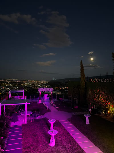 The image depicts an outdoor event space at night, likely in the Lisi, Georgia area, overlooking a city illuminated by numerous lights. The foreground is dominated by a lawn area with pathways illuminated by vibrant pink and purple lights. Several small, round tables covered with fabric are scattered across the lawn, with two tables prominently featured in the center, each with a small object on top. In the middle ground, a white structure with a canopy and seating arrangements is visible, also illuminated with pink lights. Adjacent to this is an archway decorated with white flowers. A pathway leads towards this archway.

The background reveals a dense cityscape stretching into the distance. In the upper right quadrant of the image, a sign reads "LOVE GARDEN" in illuminated letters against a dark green, possibly hedge-like, backdrop. A tall, slender tree stands to the left of the sign. A man is visible on the right side of the image, standing near some bushes and lighting equipment. The night sky above is dark blue with scattered clouds and a bright full moon. The overall atmosphere suggests a festive or romantic evening event.