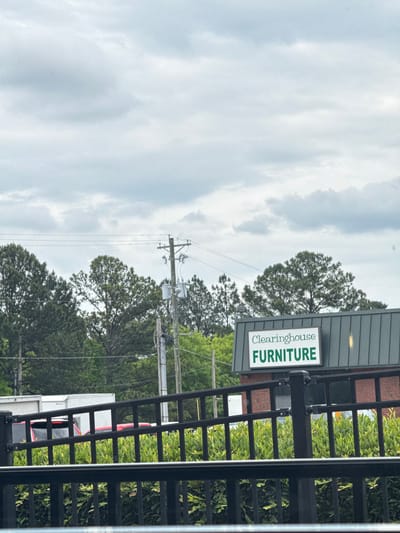 The image depicts a roadside scene near Stone Mountain, Georgia. The focal point is a sign for "Clearinghouse Furniture," mounted on a building behind a dark metal fence. Power lines and a utility pole rise in the background, partially obscured by trees. A glimpse of a white truck or trailer with red accents is visible behind the fence and foliage. The sky is overcast with heavy clouds.