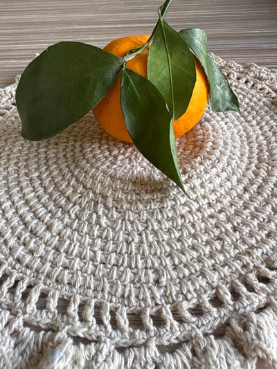 A close-up, overhead shot shows an orange with green leaves resting on a crocheted placemat. The placemat is beige and has a circular, concentric pattern with a lacy edge. The background appears to be a wooden surface. The image is a still life. The location is not identifiable within the city of Bratsk, Russia.