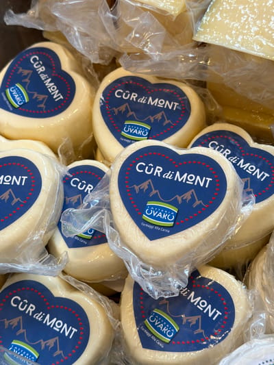 This image shows a close-up, overhead view of several individually packaged heart-shaped cheeses. The cheeses are light yellow in color and have a smooth texture. Each cheese is wrapped in clear plastic and features a dark blue label with white lettering that reads "CUR di MONT" and "LATTERIA OVARO". The labels also depict mountains and a farm logo. The cheeses are stacked closely together, suggesting they are on display in a store or market. There are also glimpses of other cheese products and packaging in the background. The setting appears to be a food retail environment, likely in Sauris di Sopra, Italy, given the presence of the "CUR di MONT" and "LATTERIA OVARO" labels, which are associated with cheese production in that region. No people are visible in the image.