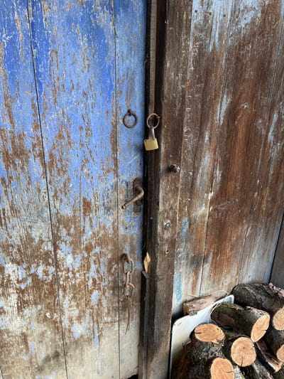 An overhead shot captures a weathered wooden surface, likely an old door or wall, in Byala Reka, Bulgaria. The surface is composed of horizontal wooden planks, with the top section a faded brown, showing considerable wear and tear. Below a prominent horizontal beam, the planks are painted a vibrant, yet heavily chipped and faded, blue, revealing the natural wood grain underneath.
Various rustic metal hardware items are affixed to the wooden surface. A brass padlock is secured through a metal ring on the left side of the central beam. To its right, a curved, rusty metal handle or latch is attached. Further right, a section of a rusty metal chain with two visible links is connected to another metal piece, extending towards the right edge of the frame.
In the upper right corner of the image, a stack of cut logs with visible growth rings is piled on a white metal tray or container. These logs appear to be firewood. The overall lighting suggests it is daytime.
On the central, weathered brown wooden plank, above the hardware, there are faint, hand-scratched markings. While largely illegible due to wear, parts of the text *might* be interpreted as "WHIT TOP OF", suggesting a possible instruction or label that has significantly faded over time. The scene evokes a rustic, outdoor setting characteristic of a rural environment.