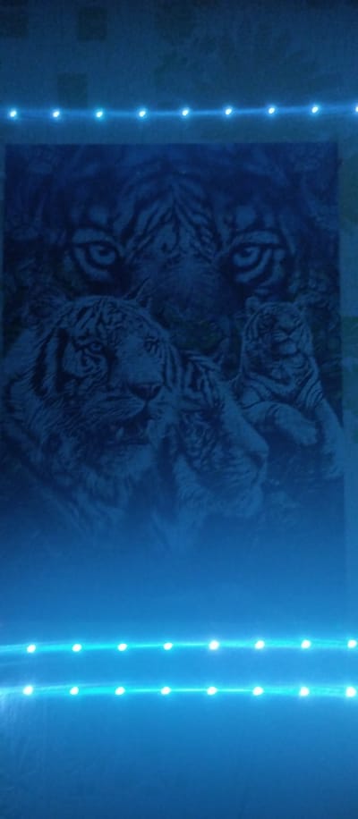 This is a vertical, medium-shot, dimly lit photograph of a painting of three tigers, likely a mother and her cubs, in a jungle setting. The painting is illuminated by blue LED strip lights along the top and bottom edges of the frame. The tigers are depicted in shades of black and white, with their stripes clearly visible. The mother tiger's face is dominant in the upper portion of the painting, with large, alert eyes. Below her, a larger cub is positioned to the left, looking forward, while a smaller cub is nestled to the right, appearing to be asleep. The background of the painting is dark and indistinct, suggesting foliage. The overall mood is one of quiet strength and maternal protection. The blue lighting casts an ethereal glow on the artwork and the surrounding wall, which appears textured. There is no visible text in the image. The location context is Aramil, Russia.