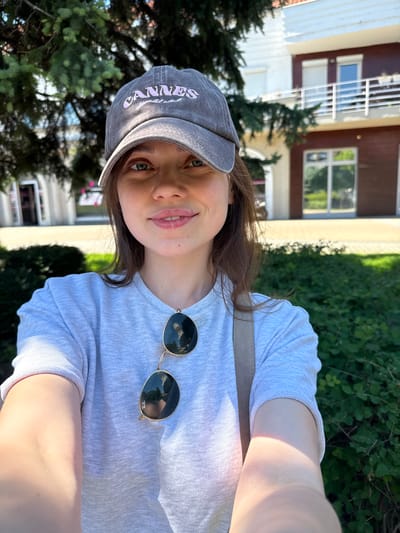 The image is a selfie of a young woman in an outdoor setting. She is wearing a grey t-shirt, a grey baseball cap with the word "CANNES" embroidered in pink, and sunglasses are hanging from her t-shirt. The background includes a building with windows and a hedge. The location is likely in the city of Siófok, Hungary.