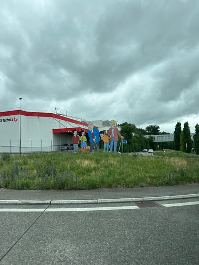 The image depicts an outdoor scene in Zurzach, Switzerland, featuring a large, flat cutout of a family, including two adults, two children, and a dog, standing in a grassy area. Behind the cutout, a white building with a red trim is visible, with the text "R Technics" and a logo on its facade. A fence runs along the front of the building. In the background, trees and a modern building with a skywalk connecting two sections are present. A blue directional sign pointing to "Basel Koblenz" is also visible. The scene is captured from the perspective of someone in a vehicle on a road, with white lane markings and a drain cover visible in the foreground. The sky is overcast with heavy clouds.