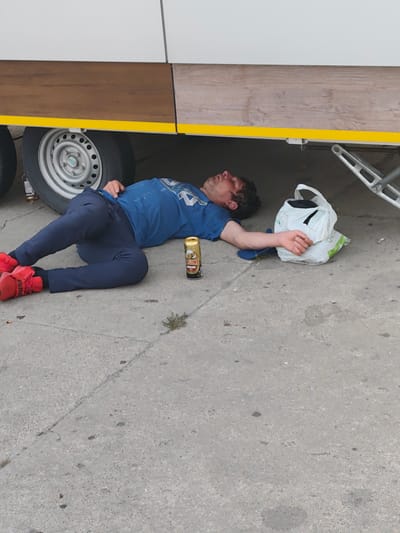 A person is lying on the ground, unconscious, under a trailer in Stryków, Poland. The person is wearing a blue t-shirt and dark blue pants, with red sneakers. A can of beer is next to the person's head. A white plastic bag is to the person's right. The ground is concrete. The trailer has a wooden-paneled side and a yellow stripe. There are two wheels visible under the trailer.