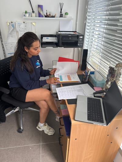 A woman is seated at a desk in an office in Maldegem, Belgium, reviewing documents. She has dark hair pulled back in a ponytail and is wearing a navy blue sweatshirt and black shorts. Her legs are crossed, and she is holding a yellow pencil in her right hand. The desk is cluttered with papers, a laptop, a printer, a tissue box, and various office supplies. The room is brightly lit by natural light coming through blinds on a window.