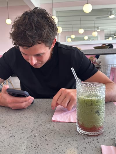 A young man in a black t-shirt is sitting at a table in a brightly lit cafe in Palm Desert, California. He is looking at his phone in his left hand while his right hand rests on a pink napkin. A tall glass of green matcha latte with a straw is on the table beside the napkin. The table is a light gray color with a speckled pattern. In the background, there are other tables and chairs, a mirror on the wall, and globe-shaped lights hanging from the ceiling.