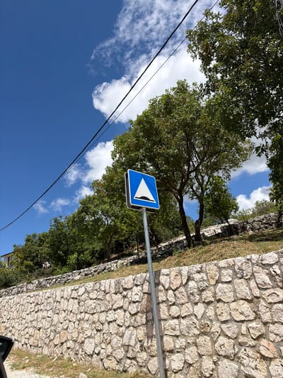 The image captures a bright, sunny daytime scene in what appears to be a rural or semi-rural setting, specifically Bratešići, Montenegro. The sky, a vibrant blue, dominates the upper and left portions of the frame, adorned with scattered, fluffy white clouds.
On the right side of the image, a robust stone wall constructed from irregularly shaped, light-colored rocks stands prominently. Patches of green grass are visible along the base and top of this wall. To the left and top, dense green foliage from various trees creates a natural canopy, with sunlight filtering through the leaves. One large tree with visible branches is particularly central on the left side of the frame.
A blue square road sign with a white border is positioned towards the center-right, mounted on a silver metallic pole. The sign features a white arrow, depicted as two chevrons, pointing diagonally upwards and to the left, indicating a directional warning or instruction, likely for a sharp bend or obstacle.
Two dark utility lines or cables stretch diagonally across the sky from the lower left to the upper right, adding a man-made element to the natural landscape. No people or activities are visible. The scene is static, highlighting the natural beauty and infrastructural elements of the location under clear, bright weather conditions. There is no discernible text on the sign, only the graphic symbol.