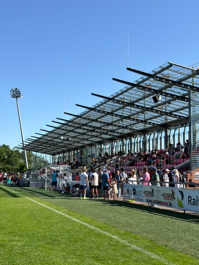 The image depicts a soccer field in Fentange, Luxembourg on a sunny day. Spectators are visible in the covered seating area, a structure with a latticework glass roof. A tall lighting tower stands on the left side of the field, under a clear blue sky. A white sideline is prominent in the foreground. Advertising banners are affixed to the seating area.