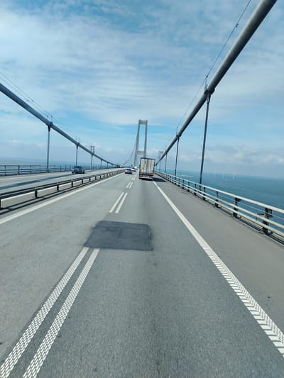 This is a POV shot from a vehicle traveling across a large bridge. The bridge spans over a body of water, and on the horizon, several wind turbines are visible. The road surface is asphalt with clear lane markings, including double white lines and textured rumble strips on the edges. The bridge has a metal railing on the right side and a large suspension cable running parallel to the road overhead, supported by vertical cables. Several other vehicles, including cars and a truck, are on the bridge. The sky is partly cloudy with a bright blue hue. The location is identified as the Great Belt Bridge in Denmark.