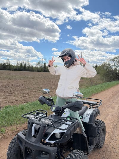 A woman in a helmet and light-colored jacket poses on an ATV in a rural area near Можга, Russia. The ATV is primarily black and white, with front and rear racks. The woman is making a peace sign with both hands. The landscape includes a plowed field, a dirt road, and patches of grass. The sky is blue with puffy white clouds, and power lines are visible.