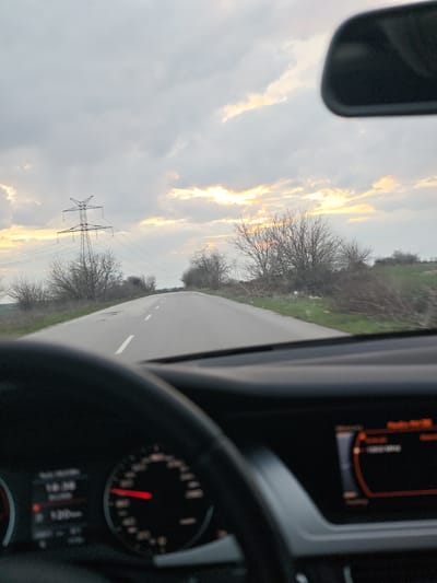 The image captures a forward perspective from inside a vehicle on a paved road located in Gorni Tsibar, Bulgaria. The time is approximately 18:00, indicated by a blurred digital display within the instrument cluster visible behind a dark steering wheel in the lower left. A red needle on the speedometer is partially discernible. A blurred infotainment screen is present on the lower right dashboard, featuring an orange-toned interface. A portion of the vehicle's rearview mirror is visible in the upper right.
The road is asphalt, marked with white dashed center lines, and extends into the distance, exhibiting a slight upward grade and gentle curve to the right. Both road shoulders are covered in green vegetation, with numerous bare deciduous trees and shrubs lining the route. On the left side of the road, a prominent metal electricity transmission tower stands, supporting multiple power lines that traverse the landscape. The sky is predominantly cloudy, with distinct areas of orange and yellow light on the horizon, consistent with twilight conditions.