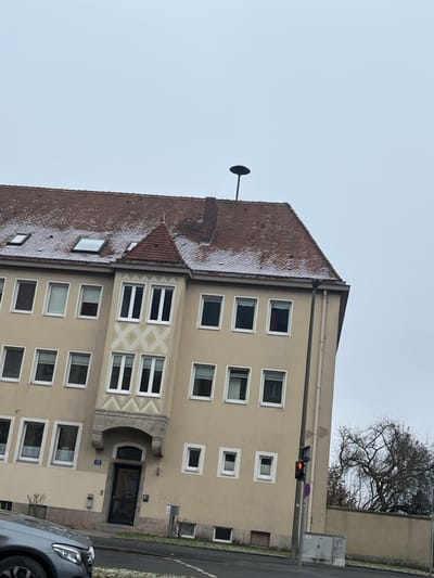 This image presents a multi-story residential building located along a street in Schweinfurt, Germany, on an overcast day. The building features a pale beige or light yellow facade with numerous white-framed rectangular windows, some of which are slightly ajar.

The reddish-brown tiled roof shows traces of light snow or frost, indicating cold weather. A distinctive feature on the roof is a large stork's nest, positioned on a pole next to a brick chimney. Several skylight windows are also visible on the roof. The central part of the facade, above and around the main entrance, is decorated with intricate geometric patterns in a lighter hue, including diamond and chevron designs. The main entrance is recessed beneath a stone archway and leads to a dark-colored door. A small blue house number plaque, appearing to display "9", is visible next to the entrance.

Adjacent to the building, a paved road stretches, with a modern gray car (a Mercedes-Benz, identifiable by its headlight and grille) parked in the foreground. A traffic light, displaying a red signal, stands on a pole near the corner of the building. In the background, behind the building, a deciduous tree with bare branches reinforces the indication of winter or late autumn. The sky is uniformly light gray and overcast. The scene is static, devoid of visible human activity, capturing a typical urban street view.