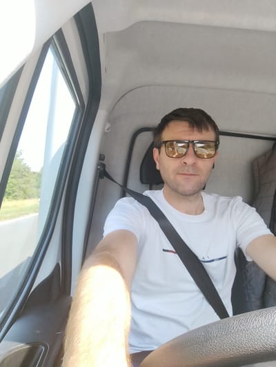 The image shows a man with short brown hair and sunglasses, wearing a white t-shirt and a seatbelt, sitting in the driver's seat of a vehicle. The interior of the vehicle is visible, including the dashboard and a portion of the steering wheel. Outside the vehicle, through the window, a grassy area with trees and a road can be seen. The scene appears to be a sunny day. The location is not specified as Kist, Germany.
