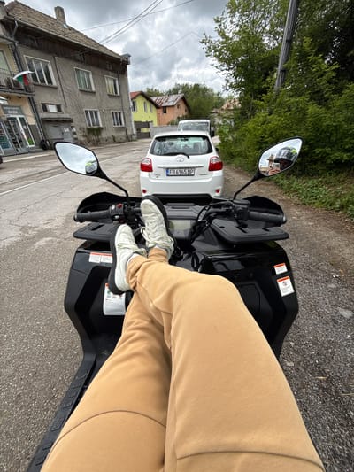 The image depicts a person seated on a black quad bike on a street in the city of Брусовeте, Bulgaria. The person is wearing light-colored pants and white sneakers, with their feet resting on the bike's front section. The quad bike is parked on a paved road next to a white Toyota Auris, which has a Bulgarian license plate number EB 6965 BP. The background shows buildings characteristic of Брусовeте, including a multi-story gray building with windows and a commercial establishment on the ground floor with the sign "МИНИ МАРКЕТ ДИДИ". Other buildings in the background include a yellow and white house with a red roof. The sky is overcast. The quad bike's mirror reflects the person holding a phone.