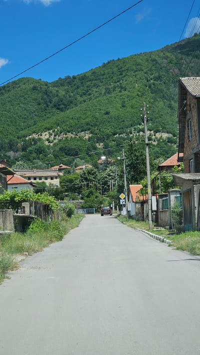 The image shows a rural road in Hristo Danovo, Bulgaria, with houses on either side and a lush green mountain in the background. The sky is a clear blue with a few wispy clouds. On the right side of the road, there is a brick building with a tiled roof, a stone wall, and a green gate. A red car is driving away from the camera down the road, which is lined with trees and utility poles. The overall impression is one of a peaceful, bucolic village setting.