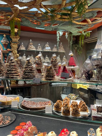 The image depicts the interior of a patisserie, specifically Pâtisserie Tonio in Sélestat, France. The scene focuses on an extensive display of confectionery and baked goods.

The upper portion of the scene features an ornate, sculptural ceiling structure resembling gnarled, intertwining branches or roots, painted in tones of gold and light grey, from which green artificial foliage hangs. An orange-toned wall section is visible above the display shelves.

Below the ceiling, a multi-tiered glass display system holds numerous cone-shaped pastries. The top two shelves are densely packed with individually plastic-wrapped pastries. Most are in the shape of fir trees (sapin), with variations including lighter-colored stacked cookies and darker, chocolate-layered versions adorned with nuts and sprinkles. Some price labels, written in French, are visible on these items, such as "Sapin croustillant" (Crispy Fir Tree) with prices for small (18€) and large (25€) sizes. On the lower glass shelf, several red, conical pastries resembling Santa hats ("Bonnet de Noël") are present, alongside a dark brown chocolate sculpture of an angel or cherub on the right.

The foreground is dominated by a glass-fronted display counter showcasing a variety of larger cakes and individual desserts. On the upper tier of this counter, from left to right, are: a large round tart or cake with a portion removed; a large round tart with a lattice top; and a tray holding multiple individual tarts or pastries topped with nuts, arranged in rows. The lower tier of the counter displays a colorful assortment of smaller, individual patisseries. These include red macarons topped with raspberries, chocolate-glazed petit fours, dome-shaped red glazed desserts, and other elaborate individual cakes with various toppings. No individuals are visible in the scene. The overall lighting is artificial and bright, illuminating the merchandise.
