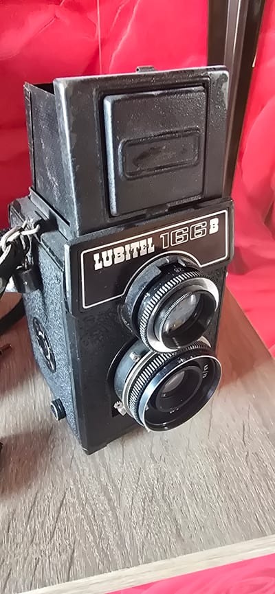 The media file shows a close-up, top-down view of a vintage Lubitel 166B twin-lens reflex camera resting on a textured, light-colored surface, possibly wood or a similar material. The camera is black and appears to be made of metal and plastic. It is positioned at an angle, with the two lenses prominently displayed. The left lens is slightly recessed, and the right lens has visible markings including "4.5/75". Silver rings are visible around the lenses, and the camera body has a textured grip.
Text visible on the camera includes "LUBITEL 166B" printed in white letters on a black rectangular plate located on the top surface, towards the left side of the lenses. There is also a smaller rectangular plate or detail on the left side of the camera body. A metal chain is attached to the bottom of the camera, suggesting it might have been used with a strap or for some other purpose.
The background is out of focus, with a vibrant red fabric on the left side and a bright pink object on the right, possibly representing a table cloth or some other surrounding material. The lighting appears to be artificial, casting subtle shadows and highlighting the textures of the camera and the surface it rests on. There are no people or animals in the image, and the scene appears to be a still life composition focused on the camera. The overall impression is one of a classic piece of photographic equipment.