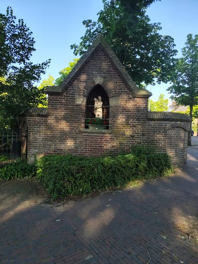 The image depicts a small, brick structure resembling a shrine or niche built into a garden wall. At the center of the structure, within an arched recess, is a statue of a woman holding a child. The statue appears to be made of a light-colored material, possibly stone or plaster. The recess is adorned with a pot of red flowers. The brickwork of the shrine and the surrounding wall is a dark reddish-brown, showing some signs of age and weathering.

The shrine is situated outdoors, with lush green bushes and plants in the foreground, partially obscuring the base of the wall. Behind the shrine, mature trees with dense green foliage are visible against a clear blue sky. The sun is casting dappled shadows on the brick wall and the paved ground in front of it, suggesting it is a sunny day. The paved area appears to be a pathway or sidewalk made of dark bricks. To the left of the shrine, a black wrought-iron fence can be seen. In the background, on the right side of the image, a white car is partially visible parked on the side of a street, and the roof of a building can be seen through the trees.

The scene evokes a sense of peace and tranquility, possibly indicating a sacred or contemplative space. The clear sky and bright sunlight suggest it is likely daytime, perhaps late morning or afternoon. The presence of the statue and the shrine points towards a religious or devotional context. The location is identified as Schaijk, Netherlands. There is no visible text in the image.