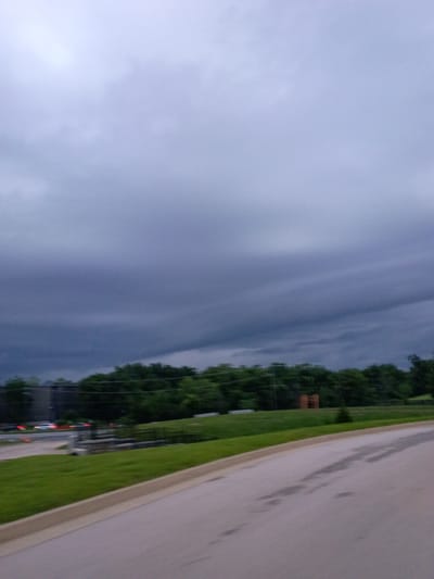 The image depicts a cloudy, overcast day in Lexington, United States of America. The scene is a roadside with a paved surface on the right, bordered by a strip of green grass. Beyond the grass, a line of lush green trees forms a backdrop, suggesting a rural or suburban setting. The sky is filled with dense, dark clouds, indicating a storm might be approaching or has recently passed. The lighting is dim, contributing to the somber mood of the image. There are no people visible, but some man-made objects are present. On the left side of the image, the sky transitions into darker, more turbulent clouds. A small, reddish-brown structure, possibly a utility box or a small shed, is visible amongst the trees. Further in the distance, faint lights from vehicles or buildings can be seen, hinting at some activity despite the overcast conditions. The overall impression is one of a quiet, somewhat melancholic natural landscape under a heavy sky.