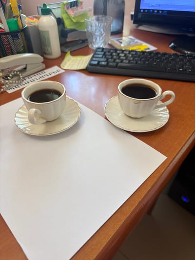 The image depicts a desk setting in Shahumian, Armenia. Two cups of coffee are prominently placed on saucers. A blank sheet of white paper occupies a significant portion of the scene, suggesting potential writing or work space. Other office supplies are visible, including a keyboard, computer monitor, pens, and a telephone. The desk surface is wooden. The lighting appears to be natural.