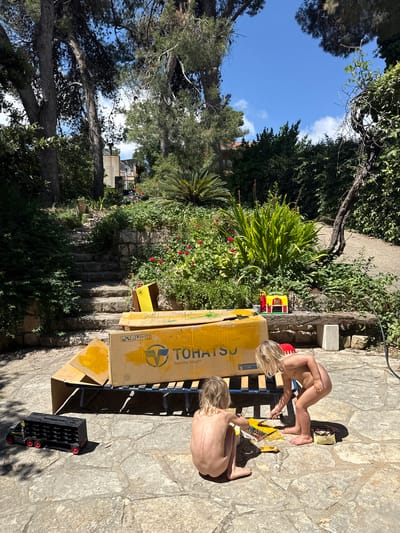 Two nude children are outside painting a box, located in Kfar Vradim, Israel. The box has the word "TOHATSU" written on it. They are sitting on a stone patio surrounded by greenery. There is a black toy truck and a yellow container near the box. A paint palette and brush are visible. The backdrop includes trees, a building, and a blue sky.
