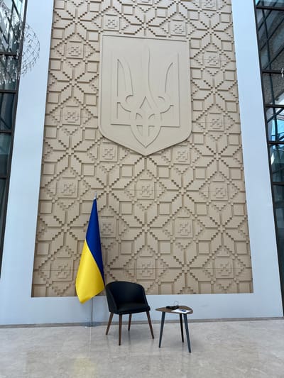 The image shows a room with a large wall featuring a decorative, embossed pattern of squares. The wall also displays the coat of arms of Ukraine, a trident, prominently centered.  A Ukrainian flag is displayed on a stand next to a black chair and a small table in the center of the room. The floor is tiled in a light color, and a portion of a large window is visible on the right side of the image. The room is located in a building at the coordinates 39.851, 32.827 in Ankara, Türkiye. The location is the Presidential Complex in Ankara, Türkiye.
