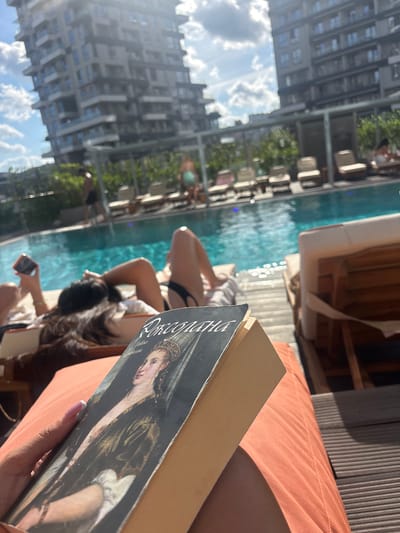 A first-person perspective shot shows a book with a painting of a woman in historical attire on the cover, resting on an orange cushion. The book's spine is visible, as is the corner of the person's hand with a pink-tipped nail, holding the book. In the background, a bright blue swimming pool reflects the sunlight, with several lounge chairs surrounding it. People are visible in the background, some near the pool and others relaxing on loungers. Two tall, modern buildings with many balconies and windows loom in the background, suggesting an urban setting. The sky is a clear blue with scattered white clouds, indicating a sunny day. The scene appears to be an outdoor poolside area in a city. The architecture of the buildings and the style of the poolside amenities suggest a contemporary urban development, possibly a hotel or residential complex in an area like Kâğıthane, Turkey, which is known for its rapid urban development and modern residential towers.