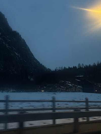 A view from a balcony shows a mountain landscape on a cloudy day. Houses are nestled on the mountainside, partially obscured by trees. Sunlight streams from the upper left corner. A railing runs along the right edge of the image, slightly blurred with water droplets. The overall scene appears to be shot during or after a light rain.
