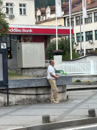 In Konjic, Bosnia and Herzegovina, a bald man in a white polo shirt and tan pants stands with his back to the camera, looking towards a fountain. He is leaning against a concrete structure in front of a building with a prominent red "Addiko Bank" sign. The fountain is spraying water, and there are yellow flowers planted nearby. The man appears to be standing idly. The background shows buildings, flags, and trees, suggesting an urban environment.