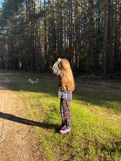 The image depicts an outdoor scene in Ust'-Silayka, Russia, within a forest environment. A single individual, a child with long, light brown hair, stands with their back mostly to the camera, facing a dense forest. The child is wearing a plaid long-sleeved shirt, dark pants with red chili pepper patterns, and purple shoes. Their right arm is raised, with the hand near their head. To the left of the child, further down a dirt path, a small, grey and white dog, possibly a Husky mix, stands on the grass, looking left.

The foreground consists of a dirt path on the left, exhibiting tire tracks and a long, distinct shadow extending from the child. The ground immediately around the child is covered in sparse green grass. The background is dominated by a thick, vertical forest composed primarily of tall pine or conifer trees. Sunlight illuminates parts of the scene, particularly the grass and the child, casting strong shadows and creating pockets of light within the darker forest. The overall environment is natural and appears to be a rural or wilderness setting.