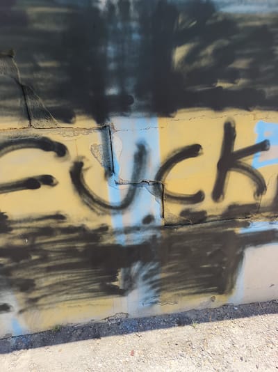 A close-up shot captures graffiti spray-painted on a concrete wall in Azor, Israel. The graffiti, rendered in black and blue spray paint on a yellow background, spells out the word "FUCK." The wall appears weathered and cracked, with some sections of paint peeling away. Darker, abstract spray-painted elements obscure parts of the text and background. The lower portion of the image shows the ground, a mix of dirt, gravel, and small pebbles, with a hint of sparse vegetation. The lighting suggests an outdoor, daytime setting.