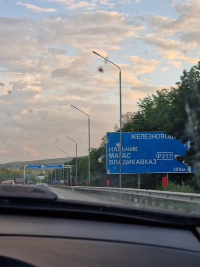 The image shows a highway scene with a large blue road sign in the foreground. The sign indicates destinations: "ЖЕЛЕЗНОВОДСК" (Zheleznovodsk) at the top, and below it "НАЛЬЧИК" (Nalchik), "МАГАС" (Magas), and "ВЛАДИКАВКАЗ" (Vladikavkaz). A route number "P217" and a distance "300M" are also visible on the sign. The road appears to be a multi-lane highway with a concrete barrier. Several streetlights are present along the roadside. In the background, there are trees and a cloudy sky. The perspective suggests the photo was taken from inside a vehicle. The presence of the town of Zmeyka is inferred by the road sign pointing to nearby cities such as Zheleznovodsk.