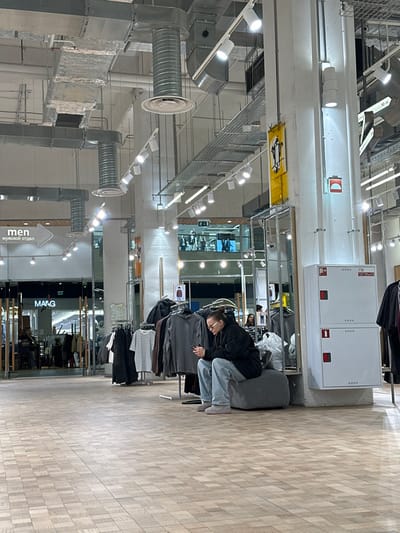 A woman in a black jacket and blue jeans sits on a gray upholstered bench in a retail store. She appears to be resting, with her head tilted slightly forward. Clothes racks filled with various garments surround her. The store has bright, overhead lighting and exposed ductwork. The "M&G" logo is visible, along with a sign indicating "men" in Russian. The floor is a light wood-grain pattern.