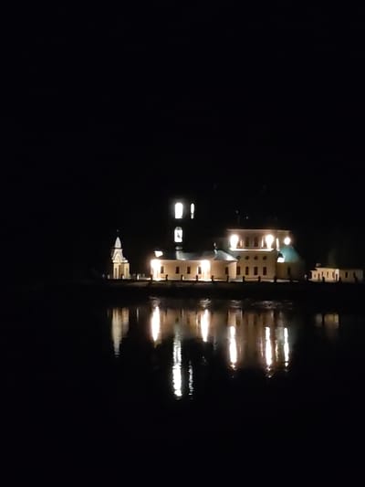 The image depicts a building at night, likely a religious structure, illuminated by artificial light. It is located in Koprino, Russia. The building has a tower and multiple sections with windows and doorways. The illumination creates reflections in the water in front of the structure. The rest of the scene is dark.