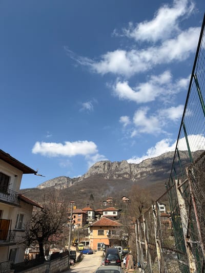 This image depicts a mountainous street scene in Gradezhnitsa, Bulgaria, under a clear blue sky with scattered white cumulus clouds.

The foreground on the left features a light-colored, two-story building with a brown tiled roof and a dark metal balcony on the second floor, along with a low stone or concrete wall bordering the street. On the right, a tall, green chain-link fence, partially obscured by bare vines and rust, runs diagonally from the mid-ground into the foreground. The mid-ground reveals a paved, curving street populated with several parked vehicles, including a dark grey hatchback, a blue sedan, and a light-colored van. A yellow excavator or construction vehicle is visible further down the street on the left. Numerous utility poles with overhead wires line the street.

The street is flanked by a cluster of multi-story houses, primarily with terracotta-tiled roofs, built into the sloping terrain. These houses display varying architectural styles and colors, including light orange and brick red. Smoke plumes are visible rising from chimneys in the mid-ground, suggesting active dwellings. The environment features numerous bare deciduous trees and bushes, indicating a dormant season, possibly late winter or early spring.

The background is dominated by large, rugged, and rocky mountains, partially covered with bare deciduous forests. Patches of shadow and light highlight the mountain's topography. No people are visible in the scene, and no specific actions or interactions are depicted.