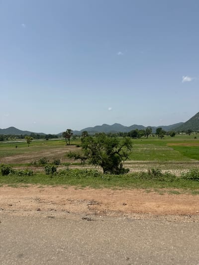 The image depicts a rural landscape under a clear blue sky with a few scattered clouds. The foreground features a dirt road with some gravel and vegetation along its edge. Beyond the road, a field stretches out, showing a mix of dry earth and green vegetation, possibly agricultural land. Several trees are scattered throughout the field, with a larger, more prominent tree in the mid-ground. In the distance, a river or body of water can be seen, flanked by more trees and rolling hills that fade into the horizon. The overall scene suggests a natural, possibly agricultural, environment in a rural area. No specific landmarks or structures that would indicate the exact location within Lankoviri, Nigeria, are visible.