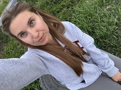 A young woman is pictured from an overhead perspective, likely a selfie. She has fair skin, light brown hair, and striking blue eyes. She is wearing a light grey hoodie with a partial view of a black and red detail on the chest. The woman is sitting on the ground in what appears to be a grassy field with patches of green grass visible. In the upper part of the image, there is a portion of a tire, suggesting she may be near a vehicle. Based on the provided information, this photograph could be taken in the city of Druzhino, Russia.