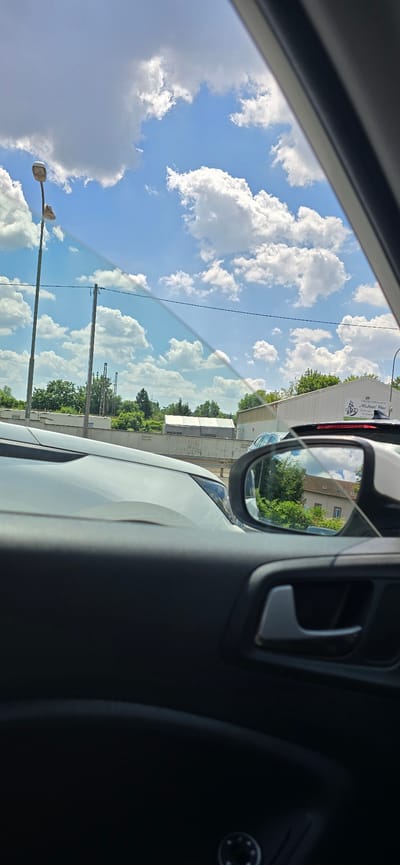 A side-view mirror reflects a building and trees. The original image shows a view from inside a car, looking out through the windshield and side window. A white car is in the foreground. In the distance, there are buildings, trees, and a bright blue sky with fluffy white clouds. There are also utility poles with wires visible against the sky. One building in the distance has a sign that reads "Steinbildhauerei Michael Hau." The scene appears to be in an industrial or semi-rural area. There are no people clearly visible in the image. The overall impression is a sunny day, possibly in Germany, given the text on the sign.
