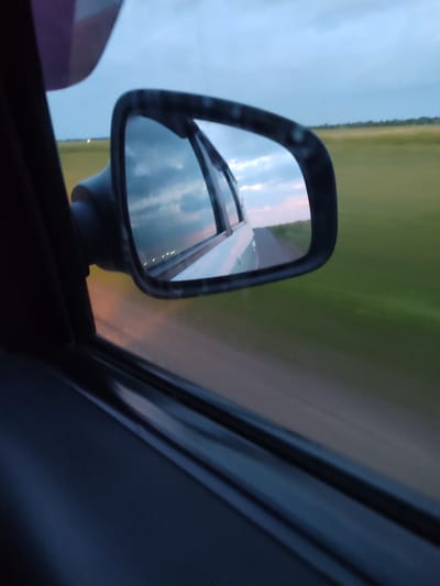 The image shows a view from the inside of a car, looking out the side window towards a wing mirror. The wing mirror reflects a scene of a road stretching into the distance, bordered by green fields on either side. The sky in the reflection is cloudy, with hints of pink and orange suggesting either sunrise or sunset.

The foreground of the image is dominated by the car's interior, with the dark interior panel of the door visible on the left. The wing mirror itself is mounted on a black arm. The background, seen through the window to the right of the mirror, is a blur of green and brown, indicating the car is in motion, likely on a rural road. The sky above the fields is a pale blue, with some clouds.

The overall impression is one of travel, perhaps on a journey through the countryside. The lighting suggests it is either early morning or late evening. There is no visible text in the image.