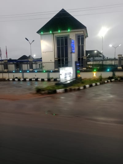 The image depicts an exterior view of a building, likely a business or commercial establishment, in the city of Kwale, Nigeria. The building has a distinctive pitched roof with green lighting along its eaves. The facade features large, dark-tinted windows and a prominent vertical sign with blue illuminated lettering that appears to display text or a logo. In front of the building, a black and white striped barrier delineates a walkway or service area from the road. The ground is wet, indicating recent rain, with reflections visible on the asphalt. Several streetlights are present, some with bright illumination. In the background, a fence with decorative pillars encloses the property. Two flags, one red and one possibly blue, are visible on flagpoles to the left of the main building. The overall scene suggests an urban setting under overcast or twilight conditions.