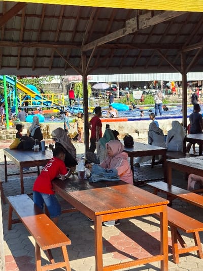 The image shows a group of people sitting and standing around tables under a large, wooden shelter in a water park in Kecamatan Tulung, Indonesia (latitude: -7.592, longitude: 110.65). A blue and yellow water slide and a small pool can be seen in the background. There is a wooden table in the foreground, with two people sitting on benches and a child standing nearby. A woman is seated at the table to the left of the image, wearing a pink hooded jacket. A small child is standing between two wooden benches in the foreground wearing a red t-shirt and blue jeans. Several other people are seated at tables in the background. The roof of the shelter is made of wooden planks. There are large trees and plants in the background, and a large building with a roof can be seen in the background.