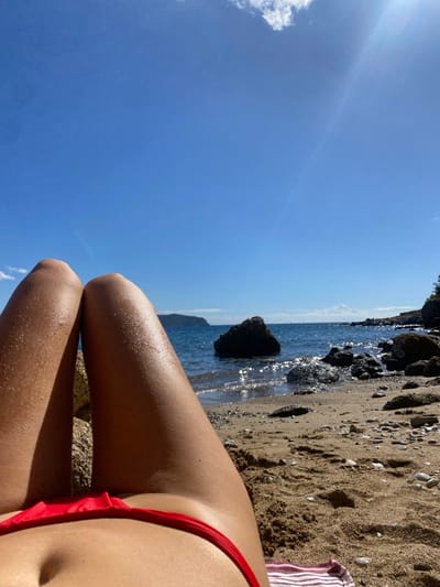 The image displays a first-person, downward view of an individual lying on a beach in Megalochori, Greece. The subject's tanned lower abdomen and upper thighs are visible in the foreground, wearing a red bikini bottom. Skin surfaces exhibit a glistening texture, suggesting moisture or oil. A red and white striped towel is partially visible beneath the right hip. The immediate environment is a beach composed of light brown sand intermixed with small pebbles and larger dark rocks. Beyond the subject's legs, the Aegean Sea extends, featuring gentle ripples and numerous sun reflections. Several dark boulders are situated both on the rocky shoreline and partially submerged in the water, with one prominent boulder centered in the mid-distance. A distant landmass is visible on the horizon. The sky is a clear blue, with a single white cloud in the upper left quadrant and a distinct sun lens flare originating from the upper right, indicating intense sunlight.