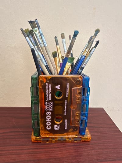 A close-up, eye-level, medium shot shows a creative collection of paintbrushes held upright in a novel pen holder crafted from repurposed cassette tapes. The pen holder is predominantly orange, with translucent green and blue cassette tape pieces forming its sides, creating a unique, retro aesthetic. The paintbrushes, of varying sizes and types, exhibit a range of bristle conditions, some appearing well-used and others newer. The bristles are a mix of natural and synthetic fibers, with some stained with paint remnants.
The cassette tape label on the orange front piece is visible, displaying Russian text and graphics, including the year "2005" and the words "САМЫЕ ЛУЧШИЕ ХИТЫ РОССИИ" (The Best Hits of Russia). The overall scene is set against a neutral, textured, pale beige wall, suggesting an indoor environment. The pen holder rests on a dark, polished wooden surface, likely a desk or table, which shows subtle wood grain patterns. The lighting is even, casting minimal shadows, and highlights the textures of the paintbrushes and the transparent plastic of the pen holder. There are no people or animals present in the image. The focus is solely on the object and its contents. The location is not specified in the image.