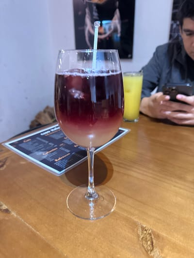 The image depicts a bar or restaurant scene, likely in Chipilo de Francisco Javier Mina (Chipilo), Mexico. A red wine cocktail sits in a wine glass with a straw, placed on top of a menu card. Adjacent is a glass containing a yellow liquid, possibly juice. A person is partially visible in the upper left corner, looking at a mobile phone. The table surface appears to be made of wood. The overall lighting suggests an indoor environment.