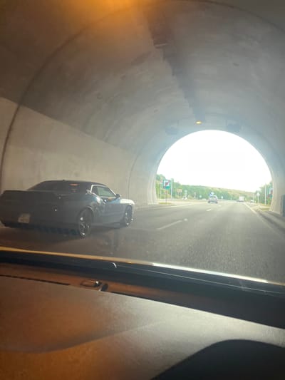 The image depicts the interior of a car, looking out through the windshield and side window, as it travels through a tunnel. On the left side of the tunnel, a dark-colored Dodge Challenger is visible, with its rear facing the camera. The car has a license plate with Cyrillic characters. The tunnel walls are made of concrete, with overhead lighting fixtures. As the car emerges from the tunnel, daylight is visible, along with a highway with other vehicles, including a white SUV in the distance. The surrounding landscape appears to be hilly and green, with trees and vegetation. There are road signs visible on the right side of the highway, indicating the end of a speed limit zone. The image's metadata does not provide specific information about the location being Blatino, Bulgaria.