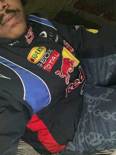 A person rests wearing a black Red Bull racing jacket with blue and red accents. The jacket features sponsor logos such as Rauch, Geox, and Total. Part of their face is visible in the corner, showing a nose ring and mustache. They are lying on a surface with a light striped pattern, with a patterned blue object behind them, possibly denim.