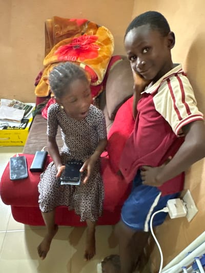 The image depicts two young children sitting on a red sofa in a room. The child on the left, a girl with braided hair, is wearing a patterned dress and looking down at a mobile phone in her hands. Next to her are two other mobile phones and a black power bank. The child on the right, a boy wearing a striped shirt and blue shorts, is leaning against the wall and looking towards the camera. A white electrical outlet with a white charging cable plugged into it is visible on the wall behind him. The room has an orange-brown painted wall and a tiled floor. A blanket with a floral pattern is draped over the back of the sofa. The overall scene appears to be indoors in a residential setting in Gidan Waziri, Nigeria.