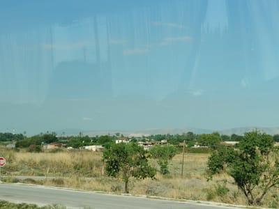 This is a medium shot captured through a window, likely from a vehicle, showing a rural landscape under a clear blue sky. The image is dominated by a reflection on the glass surface, obscuring parts of the actual scene. The reflection displays text, possibly from an advertisement or sign, reading "ISSUE DE ECO".
The visible landscape consists of dry, tall grass and sparse bushes in the foreground and middle ground. A paved road runs along the bottom left of the frame, with a red and white octagonal stop sign visible on the left side of the road. Further into the scene, a cluster of small, low-rise buildings with corrugated roofs and some solar panels are visible, suggesting a small settlement or scattered dwellings. Trees, some of which are lush green and others more sparse, are interspersed throughout the area. In the distance, rolling hills and mountains are faintly visible under a hazy sky. Two tall structures, possibly industrial smokestacks or communication towers, are present on the far left horizon.
The scene appears to be a view from the outskirts of a rural area, possibly near a highway or main road, given the presence of the stop sign. The setting suggests a warm, possibly dry climate. There are no people or animals visible in the frame. The overall impression is one of a remote, slightly undeveloped rural area in Greece, with the reflections on the window adding a layer of abstraction to the scene.