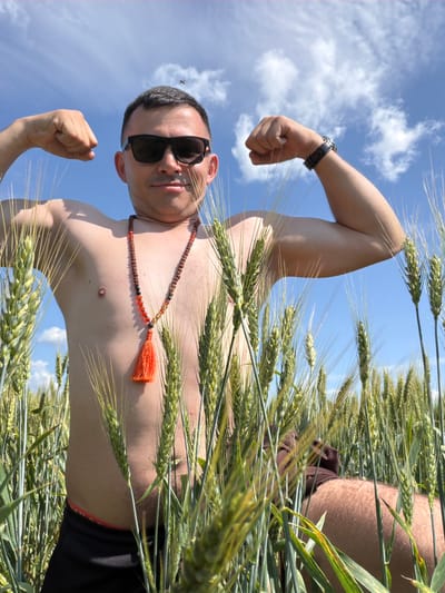 An adult male, with short dark hair and wearing sunglasses and a beaded necklace with an orange tassel, poses in a field of wheat under a bright blue sky with scattered clouds. He is shirtless, displaying his torso and arms, and is flexing his biceps. He is positioned in the foreground, with the wheat stalks obscuring his lower body. The scene is set in Podshivalovo, Russia, suggesting an outdoor, rural location. A small insect is visible in flight in the upper portion of the frame, near the sky.